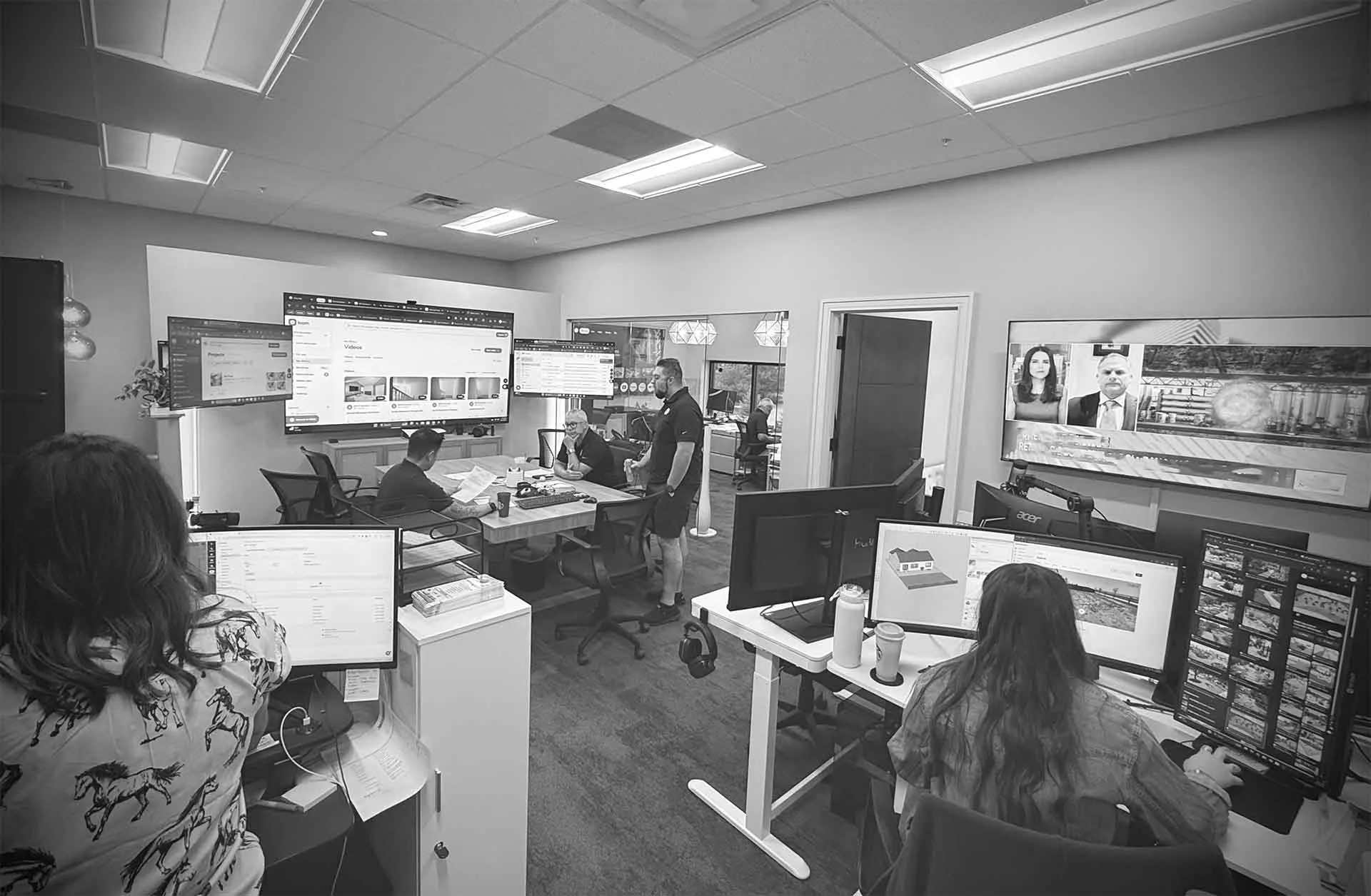 Black and white photo of a busy office environment with multiple desks and people working.