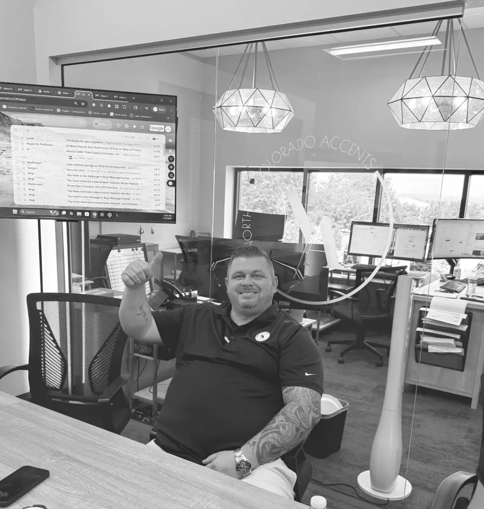 Black-and-white image of a cheerful man seated at a desk in a busy office during a team meeting.