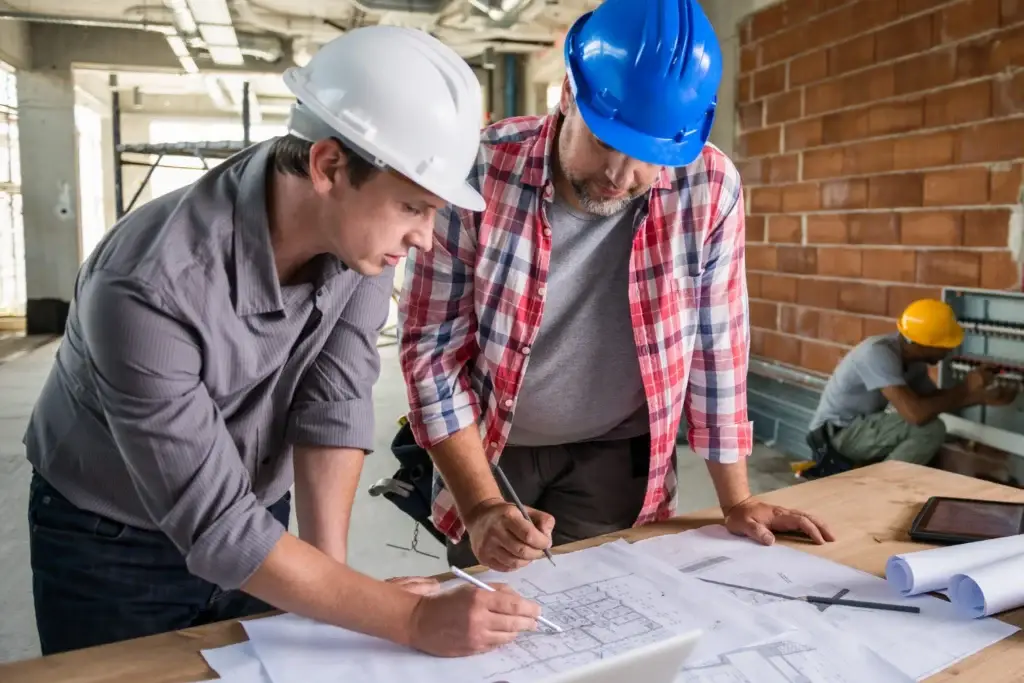 Two construction workers in hard hats reviewing blueprints on a table during a basement renovation project, with another worker installing wiring in the background.