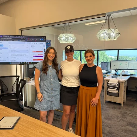 Three women posing and smiling inside a bright, modern office with computer monitors in the background.